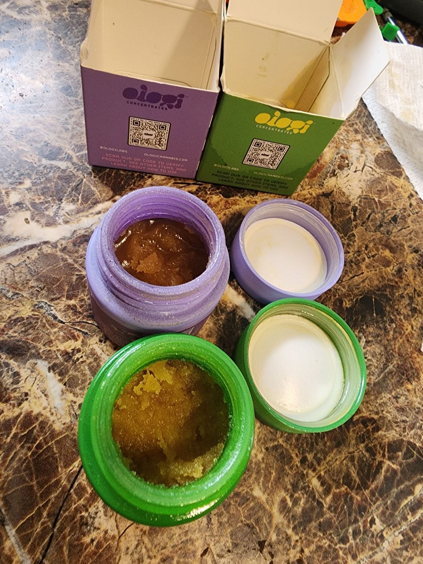 Open containers of cannabis concentrates with purple and green packaging on a marble surface.